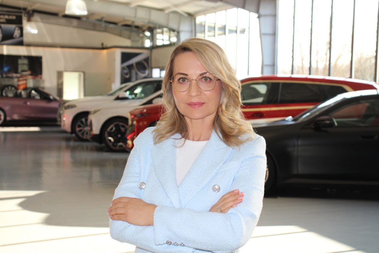 Eine Frau mit blonder Frisur und Brille steht mit verschränkten Armen in einem Autohaus